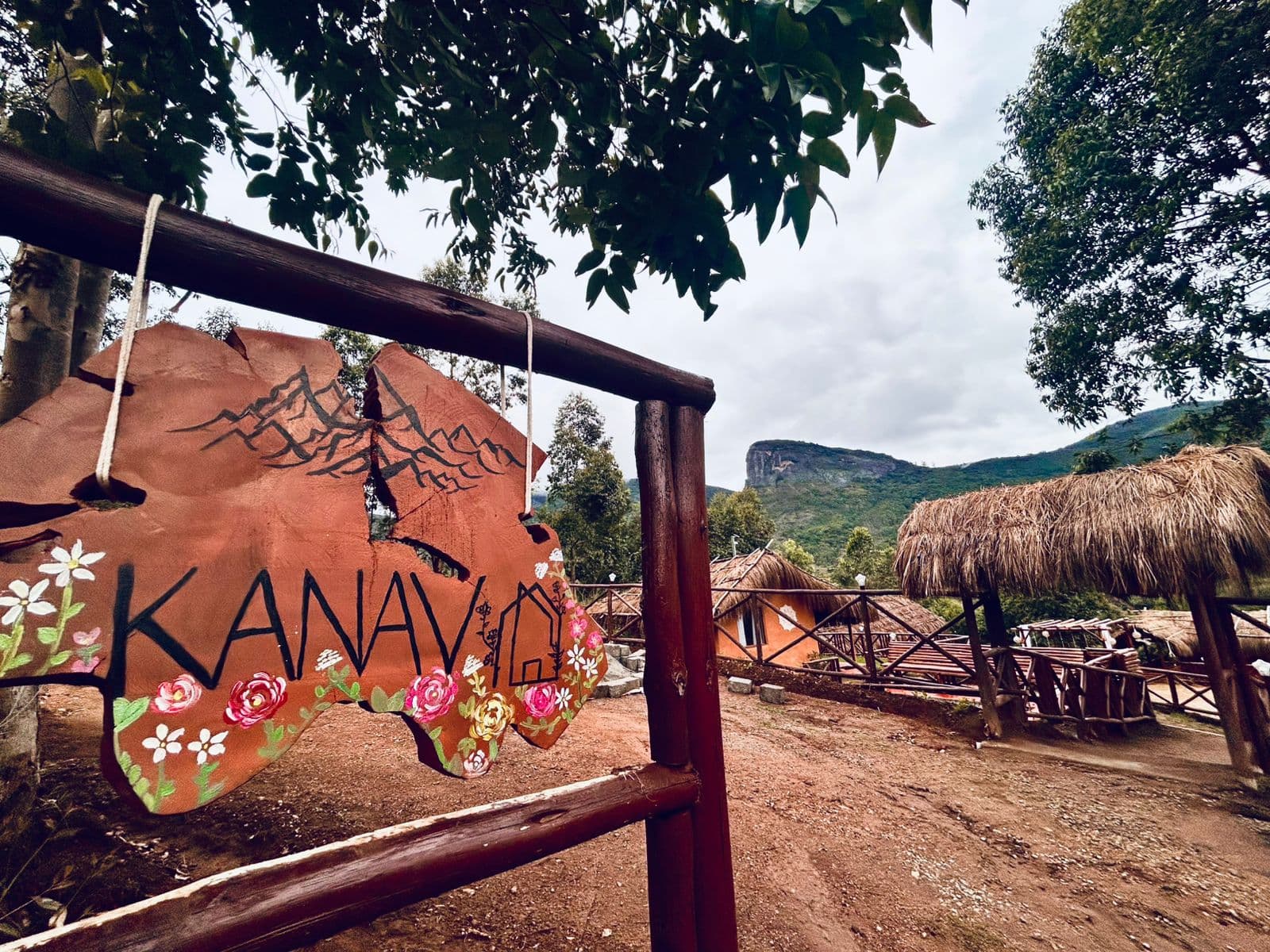 The hand-painted Kanav sign at the entrance, with thatched cottages and the mountain behind